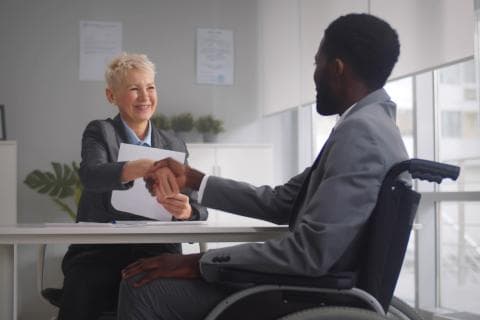 Black male job candidate in a wheelchair shaking hands with mature white woman