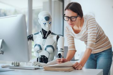 AI robot sat at a computer, while a woman in glasses sits next to pointing at a paper