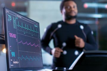 Close-up Shot of a Monitor With EKG Data. Male Athlete Runs on a Treadmill with Electrodes Attached to His Body while Sport Scientist Holds Tablet and Supervises EKG Status in the Background