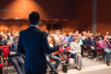 A suited man speaks at a meeting, symbolising corporatisation