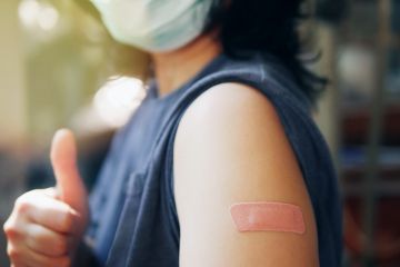 lose-up adhesive bandage on unrecognized person's arm after injection of vaccine, people in face mask received a coronavirus COVID-19 vaccine and giving thumb up to recommended inoculation