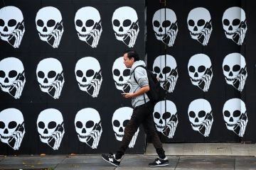 A pedestrian, using his cellphone, walks past a boarded establishment showing skeleton heads with cellphones. To illustrate that there is a delay to implementation of the lifelong learning entitlement.