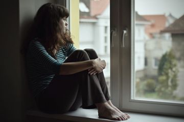 Sad young woman sitting on the window at home isolated, watching out. Coronavirus quarantine concept.