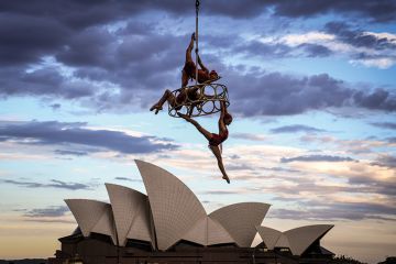 Trapeze performers hang from a cable in front of the Sydney Opera House