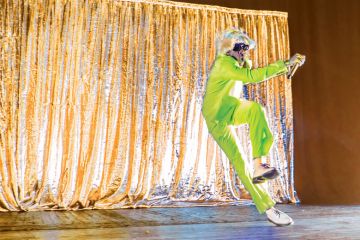 Person performing on stage, with neon green suit and blonde wig relating to scholars performing in their own online space, and being free to express themselves