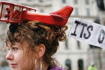 A protestor wears a toy aeroplane on her head with a banner reading  'It's over' to illustrate  ‘Set institutional carbon budgets to cut flying and support rail travel’