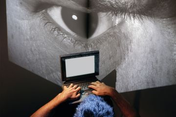  Man sits at laptop computer with image of giant eye on a big screen as a metaphor to show the online behaviour of Eric Chabriere.