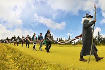 A man dressed as a grandfather leads people carrying a stick with the people holding a long knitted scarf