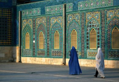 The Blue Mosque in Mazar-i-Sharif, Balkh Province in Afghanistan. Two women wearing burqas (burkas) walk past a wall of the mosque adorned with colorful tiles and mosaics. Northern Afghanistan.