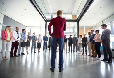 Leadership exemplified by a person in a red blazer addressing a group of diverse professionals gathered in a semicircle, in a bright modern office setting