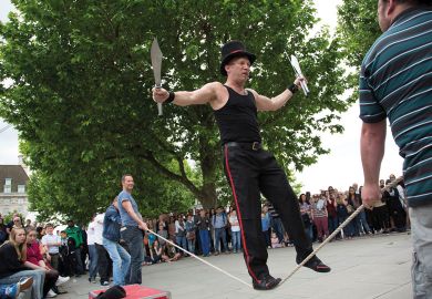 Street performer thrills the crowd with his knife act whilst balancing on a rope slack line being held up by members of the audience to illustrate UKRI ‘must regain trust’ of equality committee in Donelan row