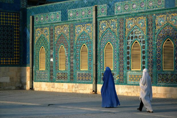 The Blue Mosque in Mazar-i-Sharif, Balkh Province in Afghanistan. Two women wearing burqas (burkas) walk past a wall of the mosque adorned with colorful tiles and mosaics. Northern Afghanistan.