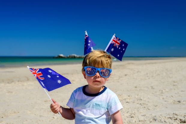A young child on a beach wearing two Australia flags on his head waves another Australia flag