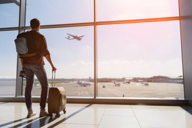 A traveller with a suitcase looks out from an airport terminal building at a plane taking off