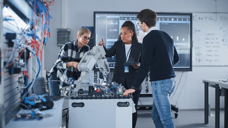 Robotics Development Laboratory: Black Female Teacher and Two Students Work With Prototype of Robotic Hand. Young Student Telling Something with Smile and Brainstorming with Her Team